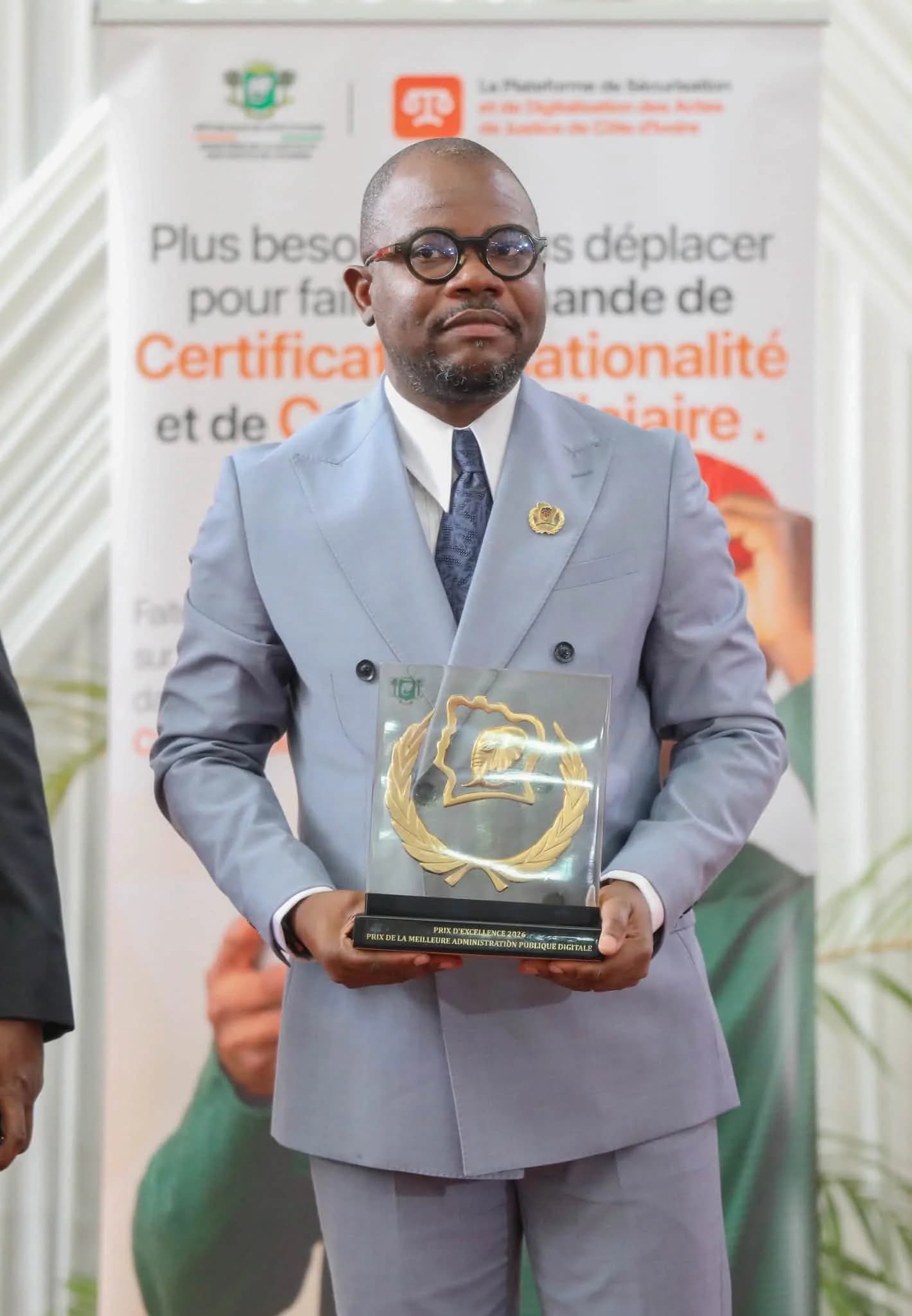 M. Auger Cadet Séoulou, coordonnateur des projets digitaux du Ministère de la Justice, présente le trophée de la meilleure Administration publique digitale devant le kakémono e-Justice